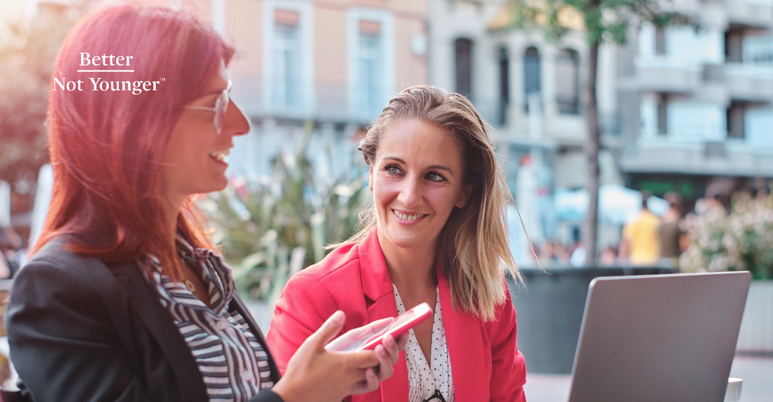 Better Not Younger features two mature women having a conversation sitting outdoors at a cafe while one of them holds a cellphone and the other one has a laptop in front of her 