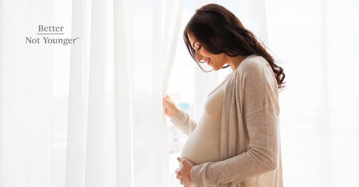 Better Not Younger features a woman who is  Becoming a First-Time Mom in her 40s. She's at home looking at her growing  belly and She's standing  by a window  with white curtains.