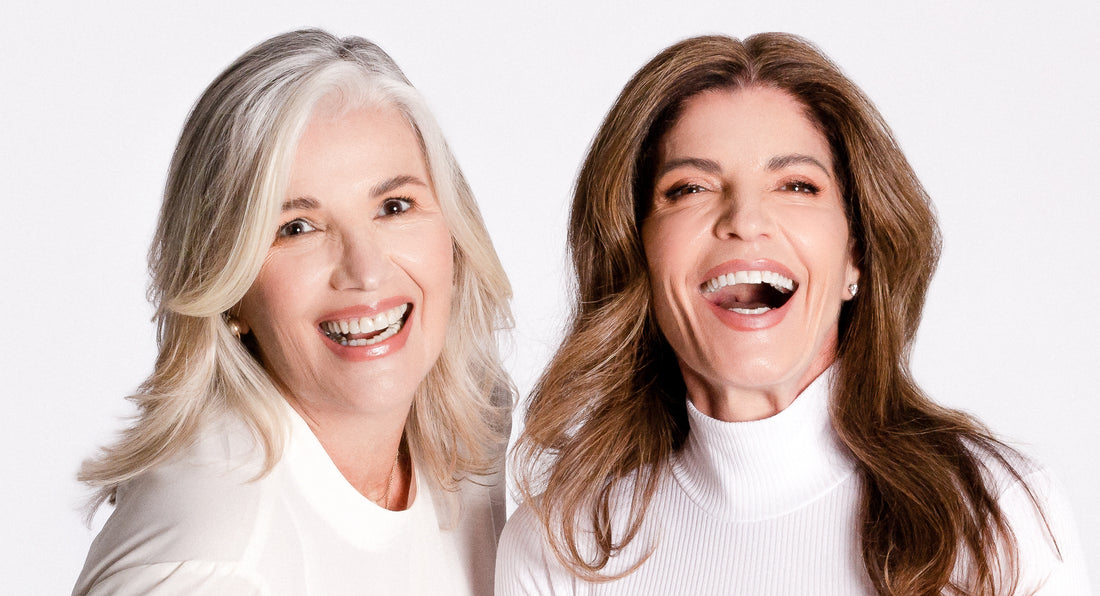 Two Women Laugh About Menopause Taboos, Both Women Look Very Happy, they wear white clothe. Woman at the left has gray long hair and woman at the right has long way brown hair. Both women are over 40 years old. 