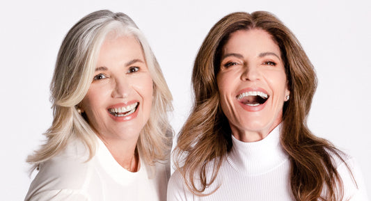 Two Women Laugh About Menopause Taboos, Both Women Look Very Happy, they wear white clothe. Woman at the left has gray long hair and woman at the right has long way brown hair. Both women are over 40 years old. 