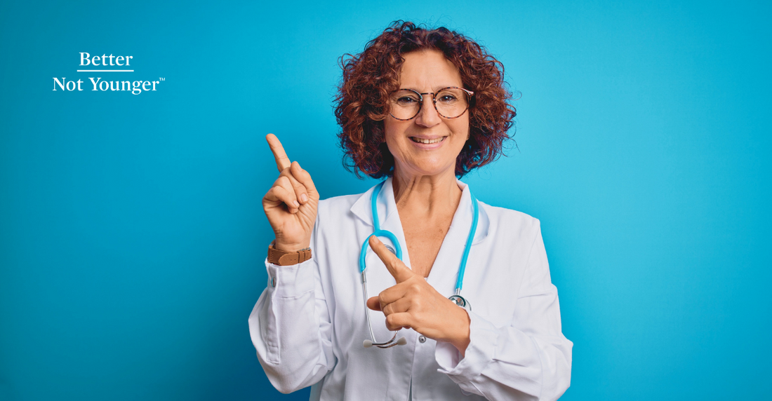 Middle age curly hair doctor woman wearing coat and stethoscope over blue background smiling and looking at the camera pointing with two hands and fingers to the Better Not Younger lettering.