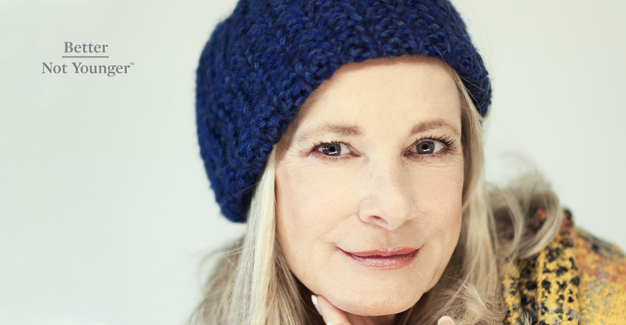 Better Not Younger features a woman over 40 wearing a blue hat hair for winter. She has blonde lng hair and she is smiling while wearing a yellow coat.