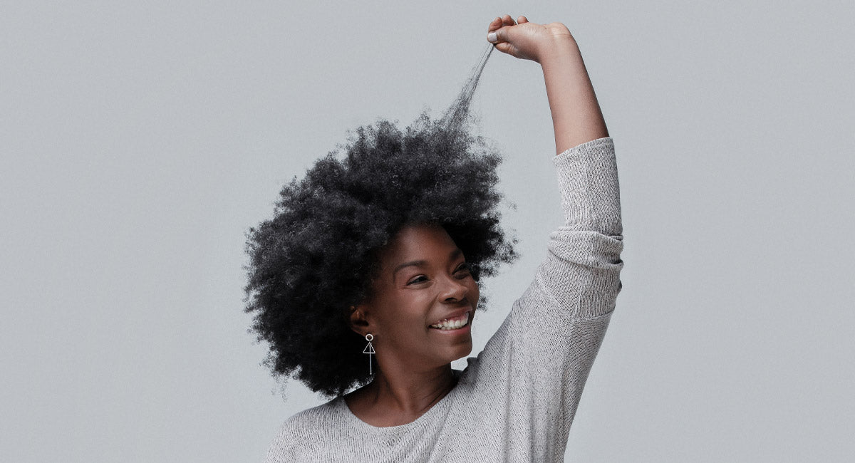 A woman holds a lock of her afro hair and looks very happy about the benefits of the Better Not Younger's hair serum she is using