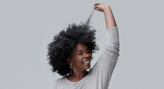 A woman holds a lock of her afro hair and looks very happy about the benefits of the Better Not Younger's hair serum she is using