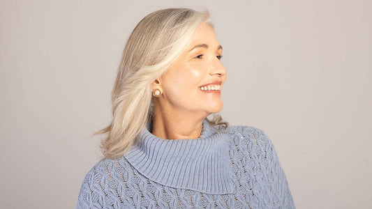 A woman over 50 feels great about her hair which is dry no more. She is wearing a purple knit sweater and has blonde medium hair. She smiles and looks to one side while we can see she's wearing beautiful luxurious earrings.