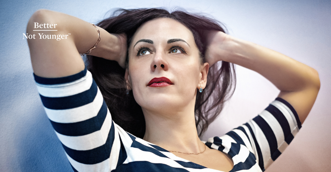 A 40 something woman with long black hair wearing black and white blouse with stripes is raising her sight to the top left corner and touching her hair