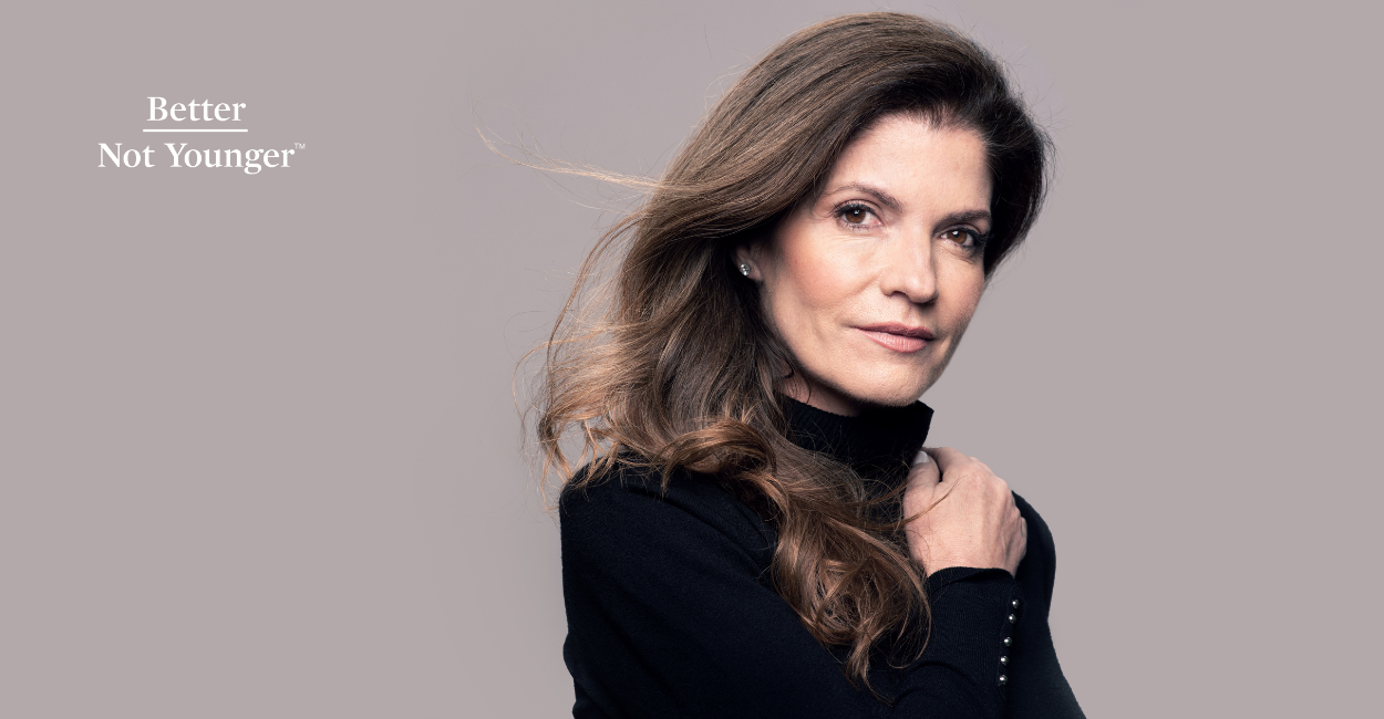 For "6 Biases Against Women’s Hair", Better Not Younger features a mature woman over 40 with long brown hair wearing a black long sleeve black blouse while her hair looks a bit blown by the air. The background is a light brown solid wall.