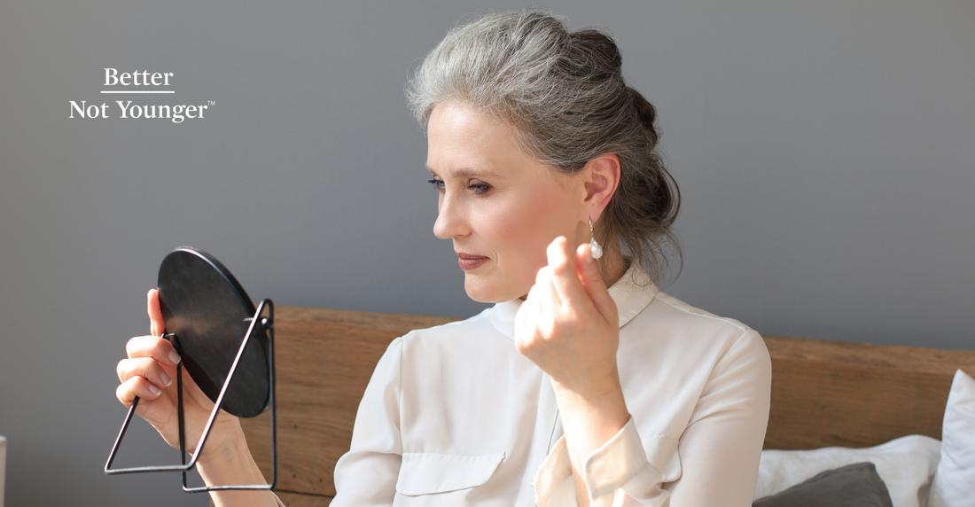 For "Why the New Hair Frosting Trend Is the Easiest Way to Grow Out Your Gray Hair", Better Not Younger features a beautiful mature woman with salt and pepper gray hair looking at herself in the mirror while wearing a white blouse with good makeup.
