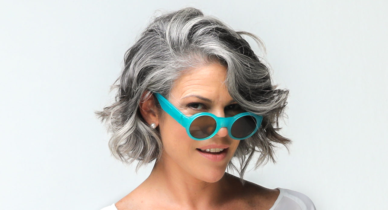 A woman over 40 years old is wearing a green long skirt, green sunglasses and holds a hat in her hands while looks sexy to the camera feeling confident about the results of her heat protection spray for her hair